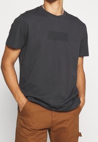 Ciemnoszara bawełniana koszulka z okrągłym dekoltem, z subtelnym wytłoczonym logo na piersi i krótkimi rękawami. Gładka tekstura i luźny krój.