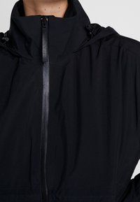 Gros plan d'une personne portant une veste noire avec un col haut et une fermeture éclair frontale avec une languette, sur un fond uni.