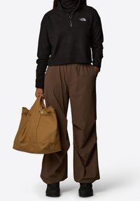 Czarny polar zapinany na zamek, brązowe spodnie szerokie z unikalnymi szwami pod kątem oraz beżowa torba na ramię z metalowymi akcentami. Całość dopełniają czarne buty.
