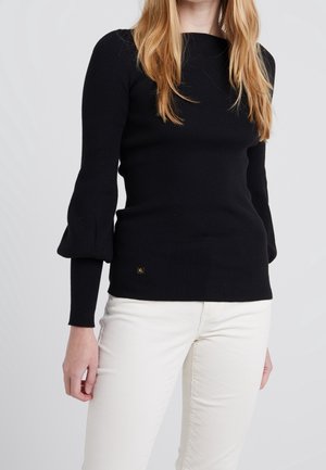 Frau trägt ein schwarzes, geripptes Langarmshirt mit Puffärmeln und weiße Hose vor schlichtem hellem Hintergrund.