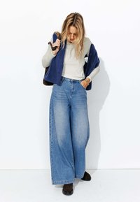Un jean bleu à jambes larges associé à un top ajust�é beige à manches longues et à un gilet en polaire marine. Des chaussures brown complètent la tenue.
