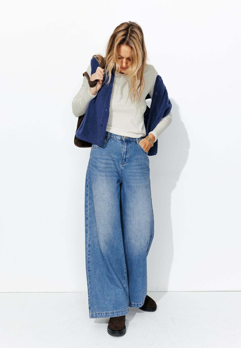 Un jean bleu à jambes larges associé à un top ajusté beige à manches longues et à un gilet en polaire marine. Des chaussures brown complètent la tenue.