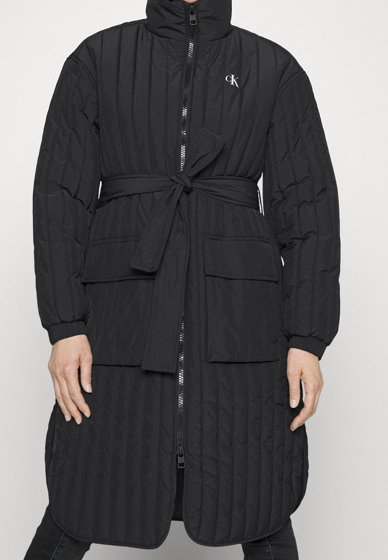 Abrigo largo negro acolchado con cuello alto, cremallera frontal, cinturón atado y dos bolsillos de parche. Presenta puños acanalados y una textura suave.