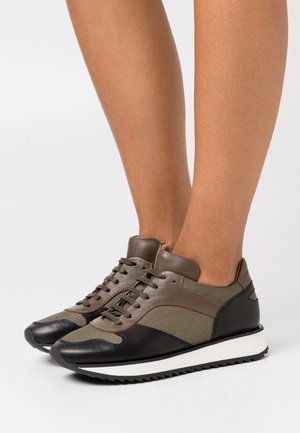 Baskets marron et noires à lacets avec semelles blanches rainurées portées pieds nus sur un fond uni.