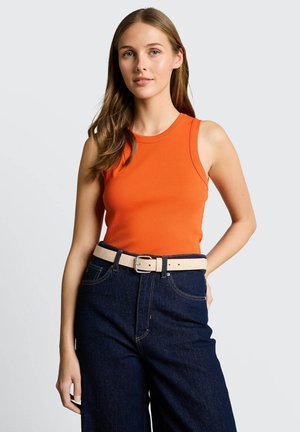 Frau mit ärmellosem orangefarbenem Top, dunkelblauen High-Waist-Jeans und beigem Gürtel, steht mit einer Hand in der Tasche vor einem hellen Hintergrund.