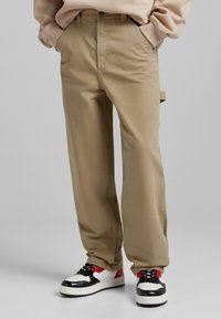 Beige puuvillahousut suoran istuvuuden, sivutaskujen ja sileän pinnan kanssa. Yhdistetty mustiin, valkoisiin ja punaisiin tennareihin.