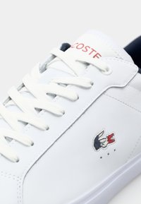 Balti odiniai sportbačiai su baltais raišteliais, ant šono su mažuoju „Lacoste“ krokodilu raudona, mėlyna ir balta spalvomis, ir lygiu paviršiumi.