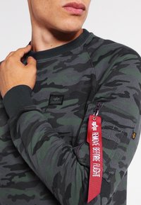 Felpa con motivo camouflage in verde scuro e grigio, realizzata in tessuto morbido. Presenta un colletto a costine e una tasca con zip con un'etichetta rossa.