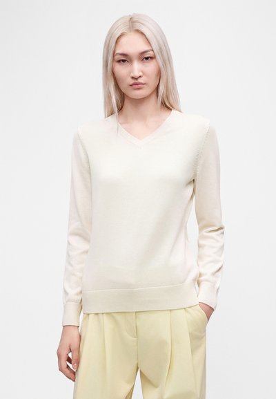 Jeune femme aux longs cheveux blonds portant un pull crème à col en V et un pantalon plissé jaune clair, debout avec une main dans la poche.