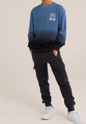 Bluza z gradientem w odcieniach niebieskiego i czarnego z logo na piersi, w połączeniu z czarnymi spodniami cargo i białymi trampkami. Styl casual, luźny krój.