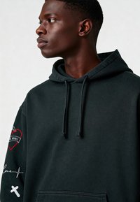 Felpa nera con cappuccio dotata di tasca frontale, cordini regolabili e dettagli ricamati, tra cui un cuore rosso e un particolare della firma.