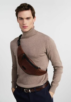 Młody mężczyzna noszący beżowy golf, ciemne spodnie oraz brązową skórzaną torbę na ramię z kieszenią na zamek.