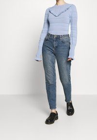 Hellblaues gestricktes Sweatshirt mit Rüschenausschnitt, langen Ärmeln und detaillierter Textur; kombiniert mit hochtaillierten blauen Jeans und schwarzen Schuhen.