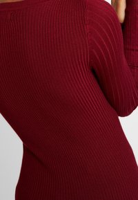 Pull rouge côtelé avec des manches longues et une coupe ajustée, mettant en valeur un tissu texturé avec des rayures verticales et une finition douce.