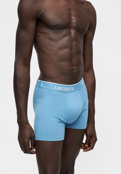 Homme torse nu portant un boxer Lacoste bleu clair avec une ceinture blanche, debout devant un fond blanc uni.