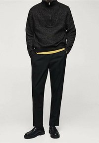 Schwarzer Strickpullover mit einem Reißverschlusskragen, kombiniert mit schwarzen, taillierten Hosen. Gelbe Schicht, die unter dem Pullover sichtbar ist. Schwarze Slipper runden den Look ab.