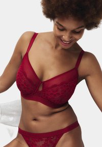 Frau lächelt und schaut nach unten, trägt ein passendes rotes Spitzen-BH- und Unterwäscheset mit Blumenmustern vor einem einfarbigen Hintergrund.
