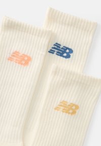 Três pares de meias creme com textura canelada, apresentando os logótipos "NB" em laranja, azul e amarelo. O design é simples e com uma textura suave.