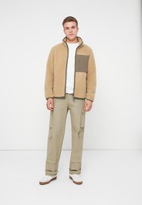 Beige Fleecejacke mit einer dunkelgrünen Brusttasche, kombiniert mit khakifarbenen Cargo-Hosen mit Seitentaschen. Darunter ein weißes T-Shirt, dazu weiße Sneaker.