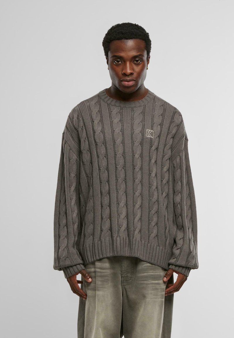 Fiatal férfi laza szürke sötétmintás pulóverben és világosszürke nadrágban, egy sima világos háttér előtt áll.