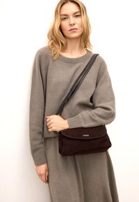 Brązowa torebka z zamszu z klapą oraz czarnym paskiem, trzymana przez osobę ubraną w jasnoszary sweter i spódnicę. Prosty i funkcjonalny design.