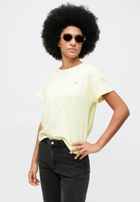 Γυναίκα με πυκνά κυματιστά μαλλιά που φοράει καφέ ηλιόμαυρα γυαλιά, κίτρινη κοντομάνικη μπλούζα και μαύρα ψηλόμεσα τζιν, ποζάροντας με το χέρι στη μέση.