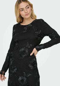 Czarna sukienka z długim rękawem, zdobiona teksturowanym kwiatowym wzorem w lekko wypukłym stylu haftu. Okrągły dekolt i dopasowana sylwetka.