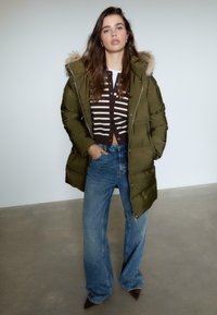 Zielona kurtka puchowa z futrzanym wykończeniem, noszona na brązowym kardiganie w paski. Dobrana z dżinsami z wysokim stanem w kolorze niebieskim oraz ciemnymi spiczastymi botkami.