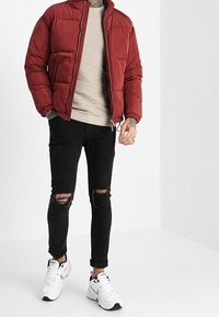 Veste rembourrée rouge, pull beige à côtes, jeans skinny noirs déchirés et baskets blanches avec des accents noirs. Matériaux texturés et coupe décontractée.