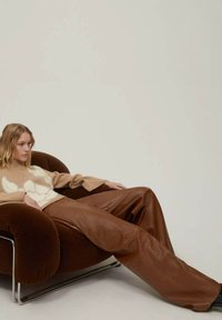Poltrona in velluto marrone con base in cromo. Modella vestita con un maglione beige con un design bianco e pantaloni di pelle marrone, seduta in una posa rilassata.