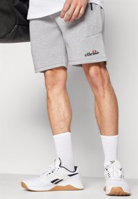 Pantalones cortos de algodón gris con un bolsillo, que presentan un logo negro de "ellesse". Llevados con calcetines blancos hasta la altura del tobillo y zapatillas deportivas blancas con detalles negros.