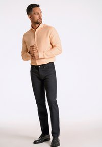 Helles oranges, knöpfbares Hemd aus einem Stoff mit feiner Struktur, kombiniert mit dunklen, schwarzen Slim-Fit-Hosen und schwarzen Schuhen.