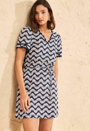 Nő sötétkék és fehér mintás, rövid ujjú, fodros szegélyű és megkötős derekú ruhában, egy texturált bézs fal előtt áll.