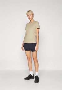 Beiges T-Shirt mit kurzen Ärmeln, kombiniert mit marineblauen Shorts. Das Model trägt schwarze Sneakers und weiße Socken und steht vor einem neutralen Hintergrund.