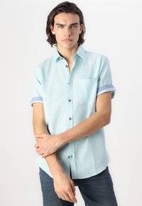 Hellblaues kurzärmliges Hemd aus gewebtem Stoff, mit einer Brusttasche und kontrastierenden blauen umgeschlagenen Ärmeln mit dunklen Knöpfen.