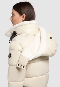 Padded cream jacket with a faux fur collar, featuring black hardware and a side button detail, textured for warmth and comfort.
