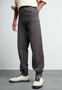 Szürke, laza fit kényelmes melegítőnadrág, ami a bokánál szűkül. Fehér Lyle & Scott logóval és oldalsó zsebekkel rendelkezik. Fehér sportcipővel viselve.