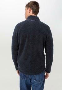 Dunkelgrauer Fleece-Pullover mit hohem Kragen und Raglanärmeln. Strukturierte Oberfläche und abgerundeter Saum, kombiniert mit blauen Jeans.