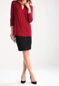 Bordeaux lange mouwen top met een V-hals en geplooid detail, gecombineerd met een zwarte kokerrok. Zwarte hoge hakschoenen en een zilveren clutch.
