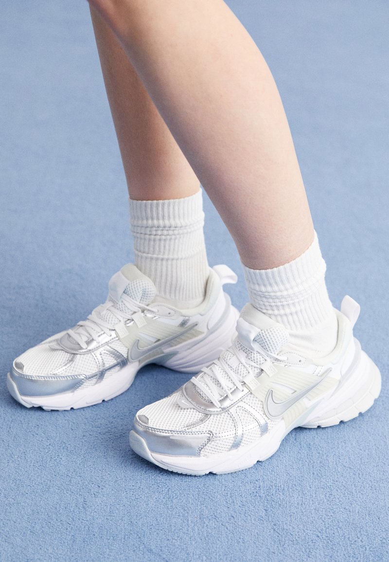Scarpe sportive bianche con sovrapposizioni in rete e sintetico, con un logo Nike argentato, abbinate a calzini bianchi a coste su una superficie blu.