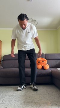 Jouet octopus en peluche orange sur un canapé gris. Une personne en T-shirt blanc et pantalon noir se tient à proximité. Des baskets noir et blanc sont au sol.
