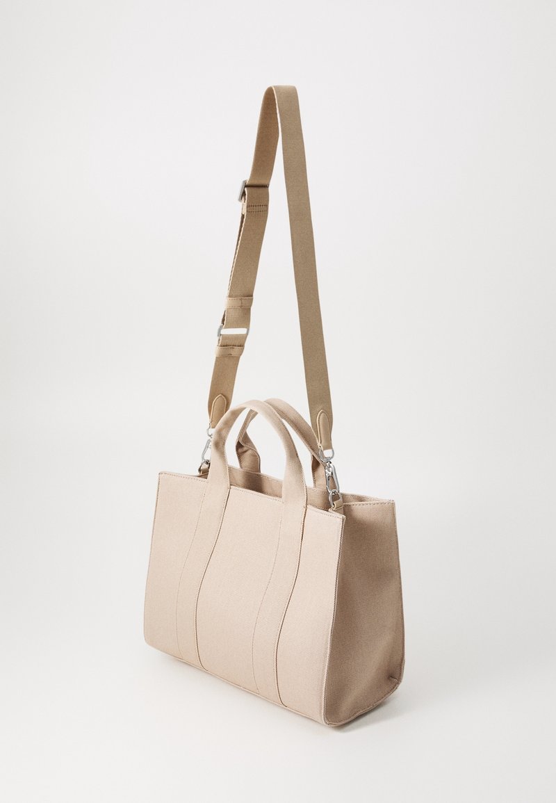 Sac fourre-tout rectangulaire en toile beige avec des poignées courtes et une longue bandoulière réglable et amovible.