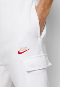 Nära bild på en hand i fickan på vita cargobyxor med en broderad röd Nike-logga ovanför ficklocket.