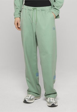 Osoba w luźnych, jasnozielonych spodniach z niebieskimi paskami przy kostkach, srebrnych butach do biegania i z ręką w kieszeni.
