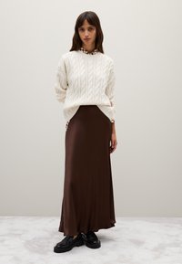 Cremfarbener Kabelstrickpullover über einem dunkelbraunen satinlangen Rock. Klobige schwarze Schuhe. Glatte Textur und schichtiger Look vor einem neutralen Hintergrund.