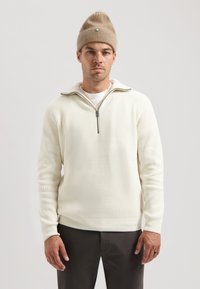 Pulover alb cu textură, cu guler cu jumătate de fermoar și mâneci lungi, purtat cu o căciulă tricotată bej și pantaloni închiși la culoare.