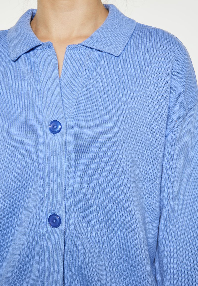 Cardigan blu in maglia con texture a coste, dotato di colletto e due grandi bottoni blu sul davanti. Materiale morbido e leggero.