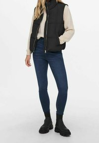 Chaleco acolchado negro sobre un suéter beige claro, combinado con jeans skinny azul oscuro y botas negras de tobillo con suela gruesa.