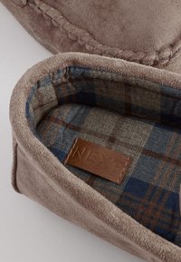 Brauner Wildleder-Hausschuh mit einer abgesteppten Kante, mit einem blau-grau karierten Futter und einem Lederpatch mit der Aufschrift "NEXT". Weiche Textur insgesamt.