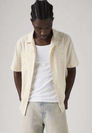 Νεαρός άνδρας με πλεξουδάκια που κοιτάζει προς τα κάτω, φορώντας ένα κρεμ πουκάμισο με κοντά μανίκια και κουμπιά πάνω από λευκό ριμπαντόν και τζιν ανοιχτού χρώματος.